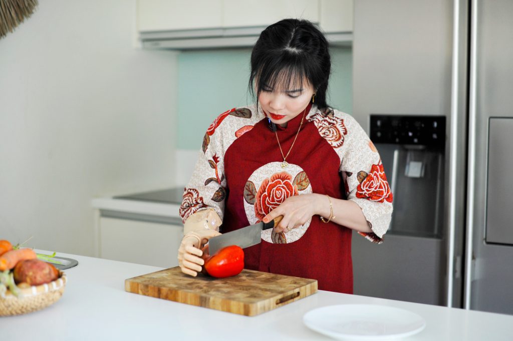 Một người phụ nữ khuyết tật tay phải đang đeo 1 cánh tay điện chức năng Vulcan để cầm ớt chuông và tay còn lại đang cầm dao cắt ớt chuông