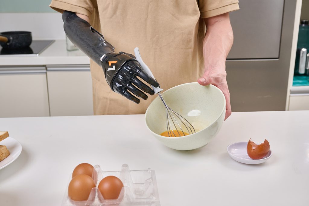 Một người đàn ông bị cắt cụt tay phải mặc áo màu be đang đeo tay giả điện chức năng Vulcan màu đen để cầm phới lồng đánh trứng trong nhà bếp