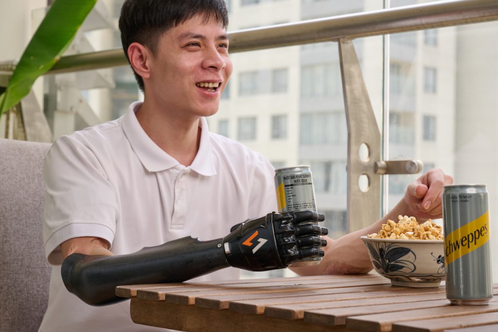 Một người đàn ông mặc áo trắng bị cắt cụt tay phải đang đeo giả điện chức năng Vulcan màu đen để cầm 1 lon bia