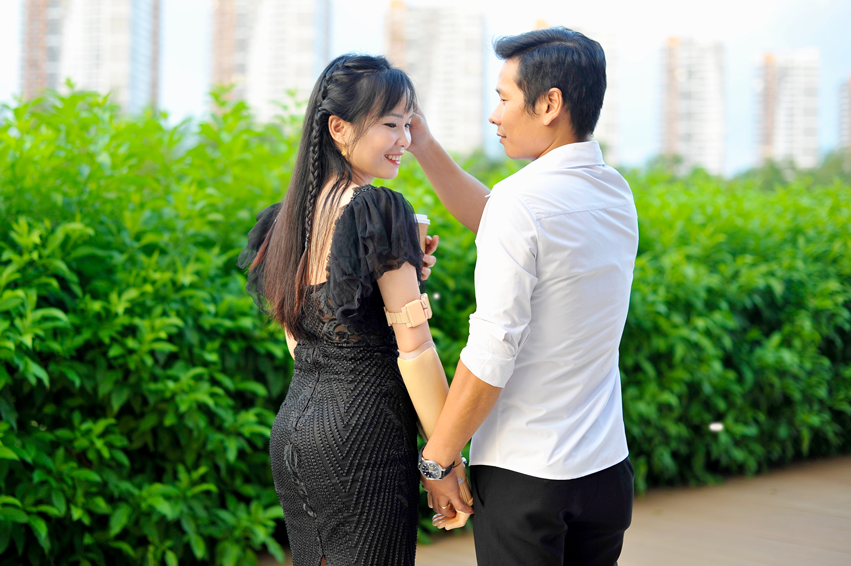 Chị Quỳnh mặc chiếc váy đen thanh lịch, đang mỉm cười hạnh phúc và nắm tay chồng dạo bước ngoài trời. Tay phải của chị đang sử dụng tay giả điện Vulcan tông màu da tự nhiên để nắm chặt lấy tay chồng, trong khi tay trái cầm một ly cà phê.