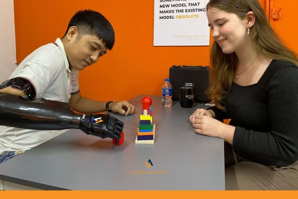 A male left hand arm amputee wearing a white polo shirt is sitting in a room and holding a lego with a woman who is wearing a black shirt