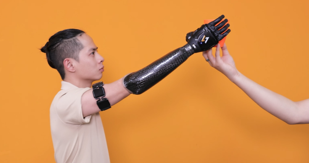 Transhumeral prosthetic user is wearing a beige shirt and the wireless Myoband. His bionic arm, featuring a carbon fiber forearm, is raised high at a steep upward angle to take an orange plastic cup from another person's hand, demonstrating high reach control against an orange-yellow background.