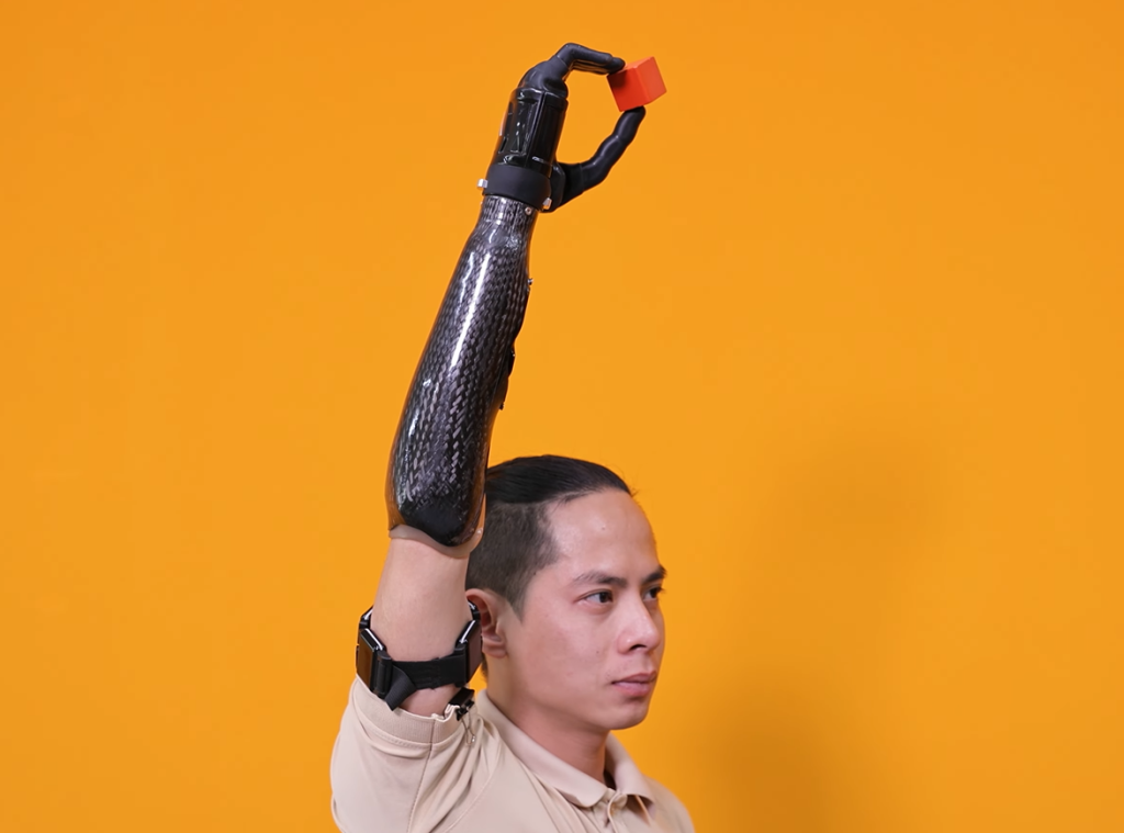 Transhumeral prosthetic user with a carbon fiber arm raises his hand high, holding a small orange block. He is wearing the black wireless multi-sensor EMG and IMU Myoband on his proximal arm against a solid orange background.